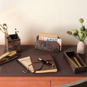 A desk with a brown notebook, pen, stapler, scissors, pencils, and two holders is complemented by the Pigeon and Poodle Chandler Blotter Set. It includes a letter organizer with envelopes and a small vase with flowers for unique workspace style.