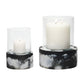 A white pillar candle burns in the Milo Black Swirled Resin Hurricane, Small: a clear glass holder with a marbled black resin base for an elegant effect.