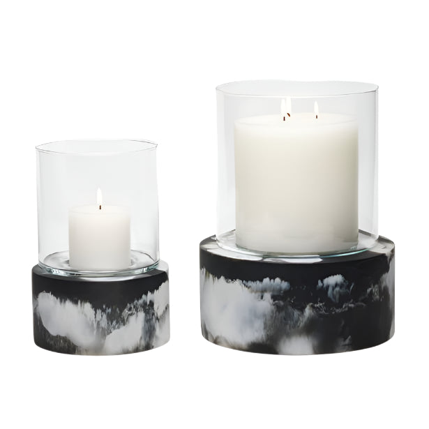 A white pillar candle burns in the Milo Black Swirled Resin Hurricane, Small: a clear glass holder with a marbled black resin base for an elegant effect.
