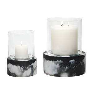A white pillar candle burns in the Milo Black Swirled Resin Hurricane, Small: a clear glass holder with a marbled black resin base for an elegant effect.