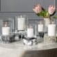 A group of white candles in large Milo Black Swirled Resin Hurricane holders rests on a round table beside a vase with pink protea flowers.