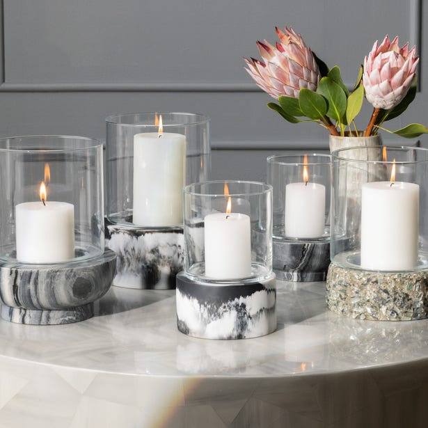 A group of white candles in large Milo Black Swirled Resin Hurricane holders rests on a round table beside a vase with pink protea flowers.