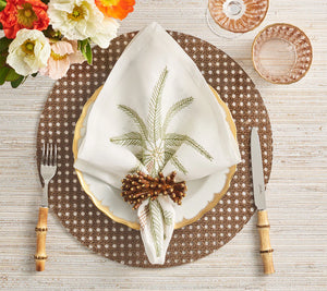 A table setting featuring the Kim Seybert Reed Placemat, a white plate with gold trim, fern-print napkin in a wooden ring, bamboo-handled cutlery, and pink glassware—perfect for stylish outdoor entertaining.