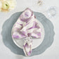 Elegant table setting featuring a white plate, lilac-patterned napkin in a Kim Seybert Cloud Napkin Ring, and a gray scalloped placemat. A wine glass and water glass are placed on the side with white flowers in a nearby vase.