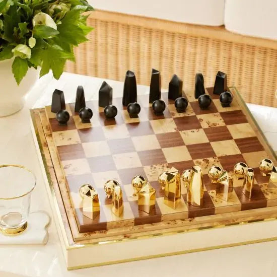 A luxurious AERIN Shagreen Chess Set featuring polished wooden squares and contemporary black and gold metallic chess pieces. A stylish game setup, completed with a glass of water and a potted plant, sits elegantly on the table.