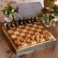 A luxurious AERIN Shagreen Chess Set with black and gold pieces is artfully displayed on a wooden table, bathed in natural light. Two small flower arrangements and a basket add charm to the scene, making it a stylish game setting.