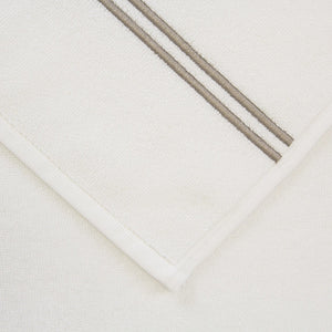 Close-up of a folded white towel with two parallel gray stripes along the edge, crafted from highly absorbent cotton terry, placed on a plain white surface. The towel is part of the Frette Classic Bath Collection, White/Khaki.