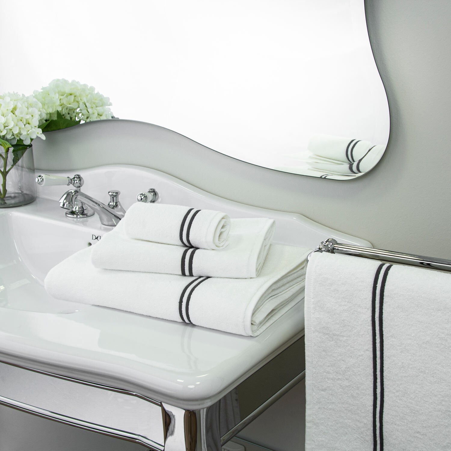 Bathroom sink with a sleek design, featuring highly absorbent cotton terry towels with double border embroidery from the Frette Classic Bath Collection, White/Grey, a chrome faucet, and a vase with white flowers. Towels are also hung on a chrome towel rack.