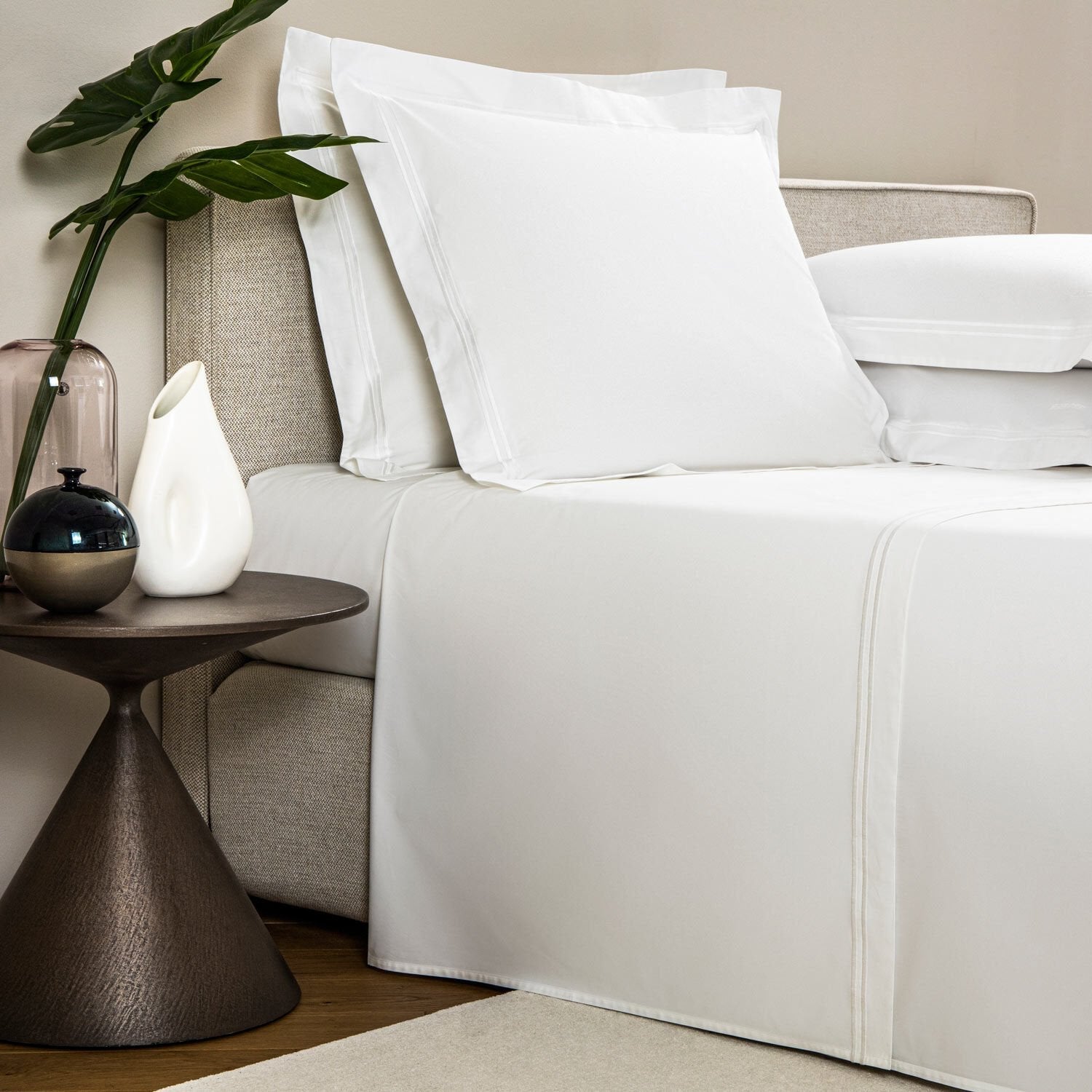 A neatly made bed with luxurious white sheets and embroidered pillows, next to a small round side table holding a white vase, a black and silver orb, and a glass container with a single leaf plant from the Frette Classic Bedding Collection, White/White.