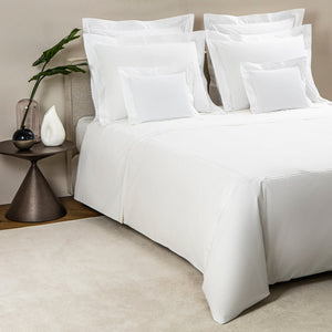 A neatly made bed with white embroidered bedding, surrounded by pillows of various sizes. A small nightstand beside the bed holds a glass vase with a green leaf and a white decorative object, exuding an aura of the Frette Classic Bedding Collection, White/White's luxurious charm.