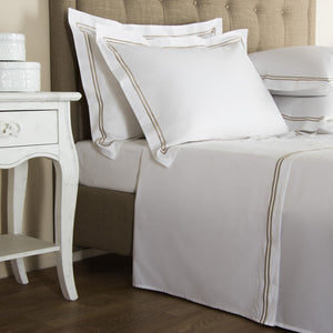 A neatly made bed with white linen and tan trim, featuring two pillows and a beige headboard. Part of our Frette Classic Bedding Collection, White/Khaki, it boasts an embroidered touch that adds to its luxurious appeal. A white nightstand with two stacked white boxes is beside the bed on a hardwood floor.