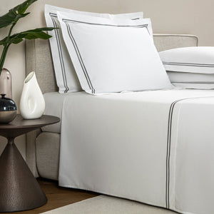 A neatly made bed with luxurious white linens and gray piping, part of our Frette Classic Bedding Collection, White/Grey, accompanied by a side table holding a white vase, a black vase, and a pink vase with a leafy plant in the background.