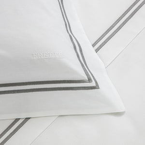 Close-up of a white pillowcase with luxurious gray double-border embroidery and the word "FRETTE" stitched in white, from the Frette Classic Bedding Collection, White/Grey.