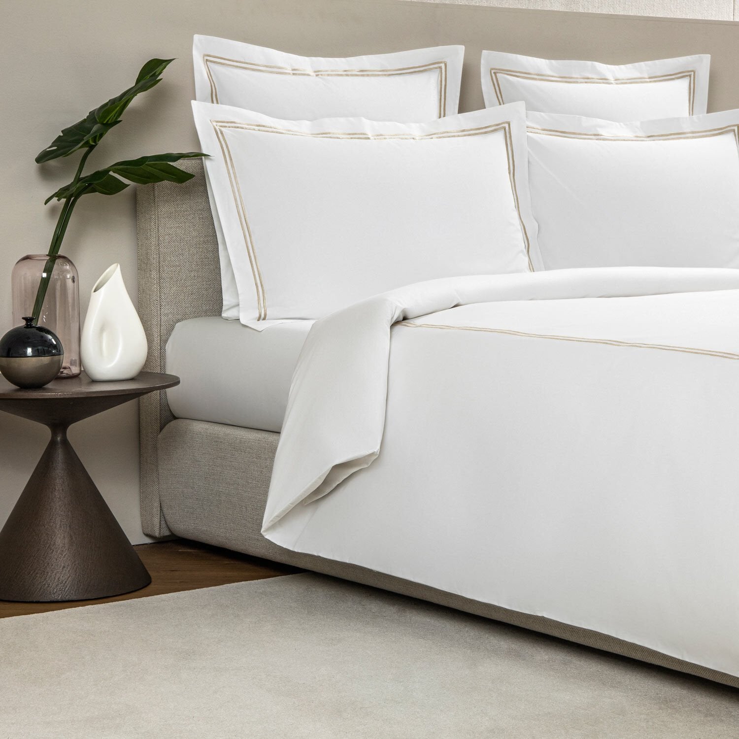 A bed with neatly arranged Frette Classic Bedding Collection, White/Khaki and multiple embroidered pillows stands beside a wooden nightstand holding a green plant, a black and white vase, and a pink vase in a modern, luxurious bedroom setting from the Classic Collection.