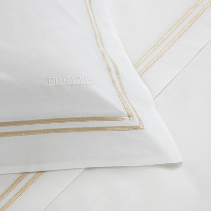 A white pillowcase and bed sheet set from the Frette Classic Bedding Collection, White/Khaki featuring luxurious gold embroidery on the borders. The pillowcase has the word "Frette" splendidly stitched on it.