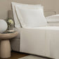 A neatly made bed with white linens from the Frette Triplo Bourdon Bedding Collection, White/Milk, featuring smooth cotton poplin and elegant three-line embroidery. The side table holds two small bowls, a vase of flowers, and a round white lamp.