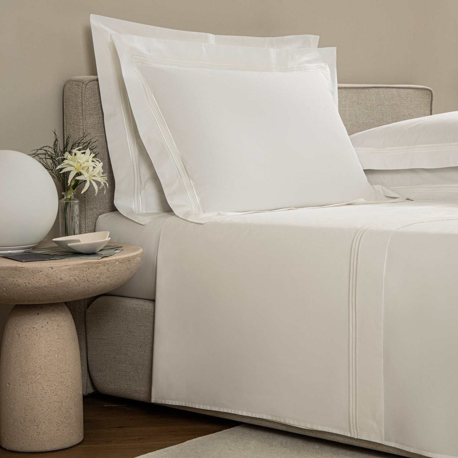 A neatly made bed with white linens from the Frette Triplo Bourdon Bedding Collection, White/Milk, featuring smooth cotton poplin and elegant three-line embroidery. The side table holds two small bowls, a vase of flowers, and a round white lamp.