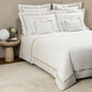 A neatly made bed with Frette Triplo Bourdon Bedding Collection in White/Beige in a tidy bedroom with neutral tones.