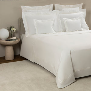 A neatly made bed with white bedding and multiple pillows featuring the elegant three-line embroidery of the Frette Triplo Bourdon Bedding Collection, White/Milk, next to a round side table holding a lamp and a small plant in a vase.