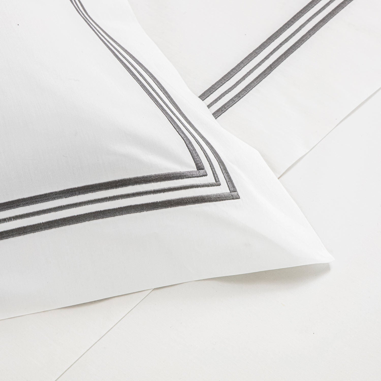 A close-up of a white pillowcase and bedsheet from the Frette Triplo Bourdon Bedding Collection, White/Grey, featuring smooth cotton poplin and two parallel black lines as a decorative border.