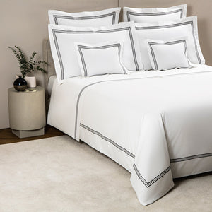 A neatly made bed from the Frette Triplo Bourdon Bedding Collection, White/Grey with smooth cotton poplin bedding featuring black trim and three-line embroidery, complemented by several matching decorative pillows. A small side table with a plant and vase sits to the left.