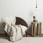 A brown chair draped with a Frette Chains Throw, Beige/Milk stands beside a round wooden side table holding two vases, a candle, and a hanging white light bulb.