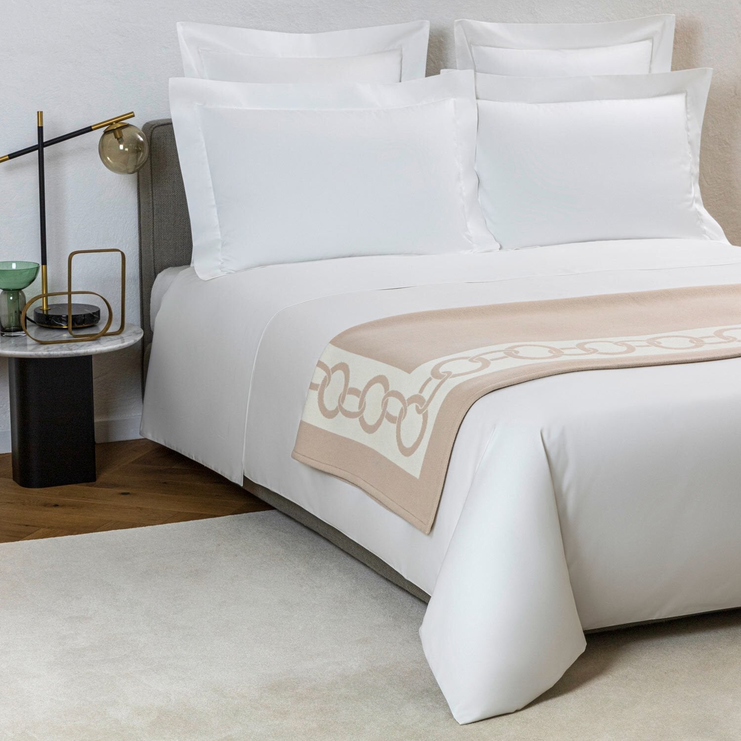 A neatly made bed with white linens, a Frette Chains Throw, Beige/Milk, pillows, a bedside table with a lamp, and a small cup. Perfect for cold winter evenings, the floor is partially covered by a rug.