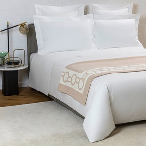 A neatly made bed with white linens, a Frette Chains Throw, Beige/Milk, pillows, a bedside table with a lamp, and a small cup. Perfect for cold winter evenings, the floor is partially covered by a rug.