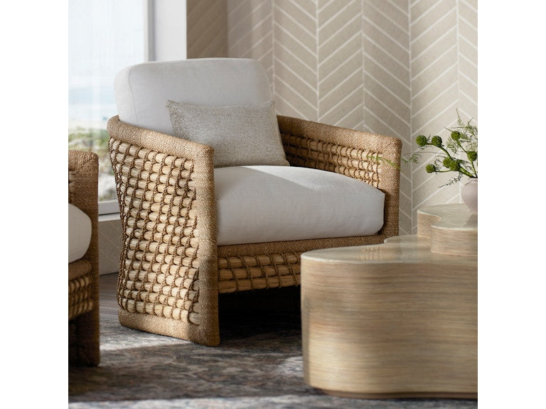The Capitola Lounge Chair, featuring woven rattan and white cushions with a small pillow, sits beside a light wood coffee table and a vase of greenery against a patterned beige wall.
