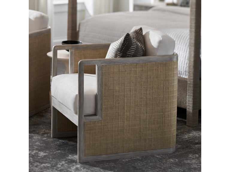 The Aveline Lounge Chair features a hardwood frame, woven side panels, and white cushions, shown on a gray rug in a bedroom.