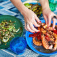 A person uses their hands to serve grilled shrimp and peppers from a blue plate on a patterned tablecloth alongside a green salad and a bottle of wine, embodying the essence of Nothing Fancy: The Art of Having People Over.