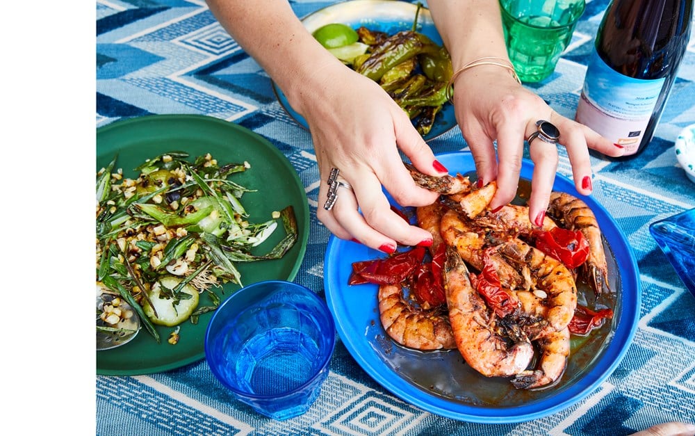 A person uses their hands to serve grilled shrimp and peppers from a blue plate on a patterned tablecloth alongside a green salad and a bottle of wine, embodying the essence of Nothing Fancy: The Art of Having People Over.