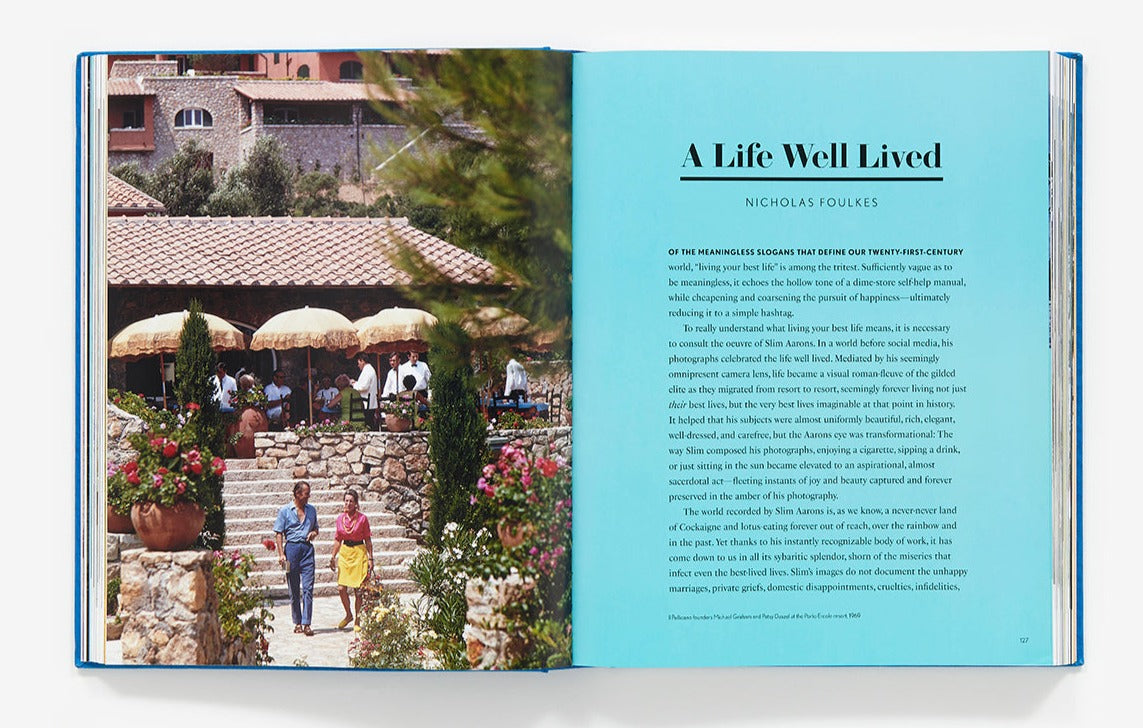 An open book titled "Slim Aarons: The Essential Collection" showcases a Mediterranean-style villa with a couple strolling on the left page, while the right page features text against a blue background. The scene evokes the elegance of Slim Aarons photography in capturing leisurely moments.