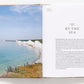 An open book titled "A Sense of Place" showcases a coastal landscape photo on the left and text "By the Sea" on the right, ideal for travel enthusiasts seeking interior design inspiration.