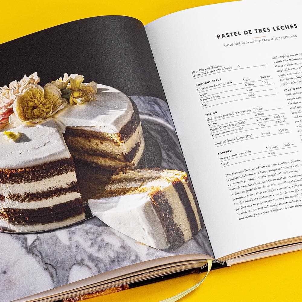 Open baking book displaying *Tartine: A Classic Revisited* with flowers on top and an adjacent recipe page detailing ingredients and instructions on a yellow background.