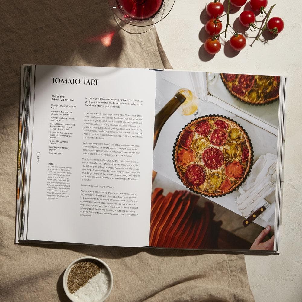 Open cookbook showing a recipe from A Table: Recipes for Cooking and Eating the French Way by Rebekah Peppler, with an overhead image of the tart alongside various ingredients on a table including cherry tomatoes, a bowl of pepper, and a glass of red wine, epitomizing the essence of French cooking.