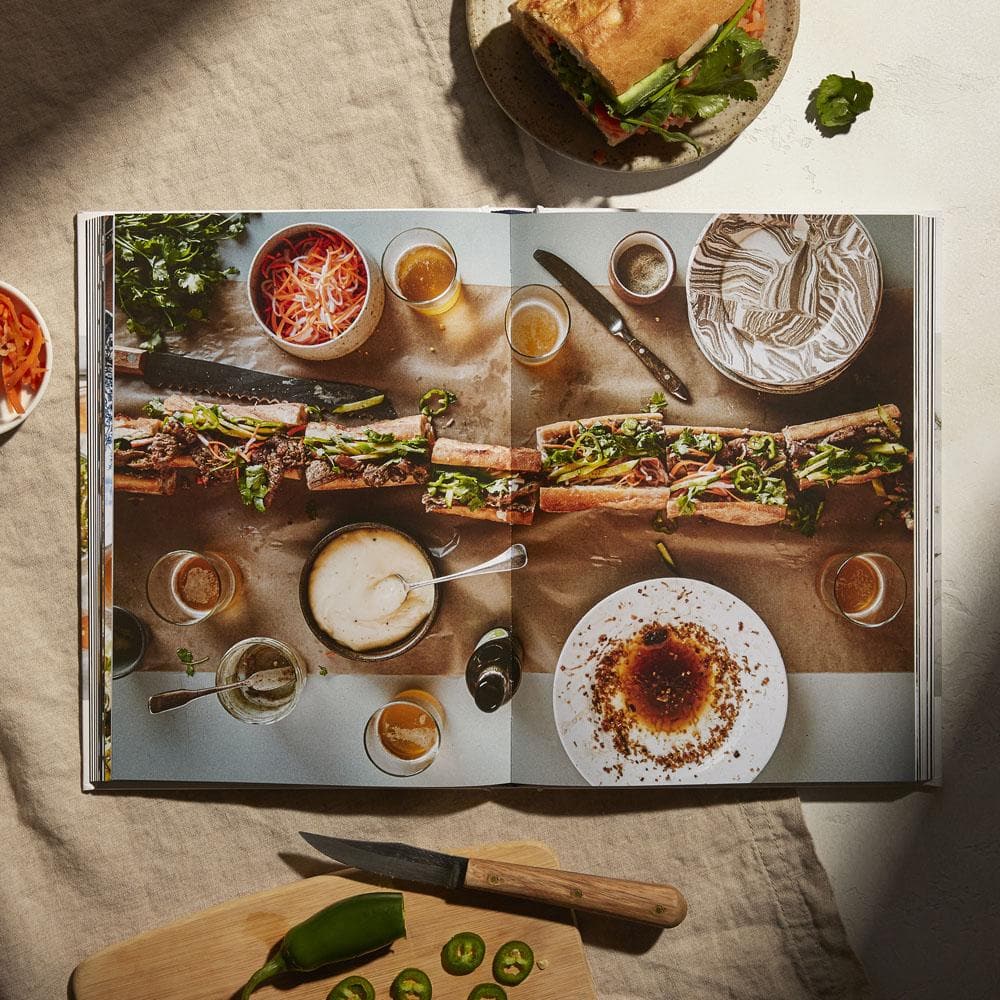 A cookbook open to a page showing a sandwich recipe. The page features a large sandwich and multiple dishes. Surrounding items include a knife, chopped vegetables, and ingredients on a table, reminiscent of *A Table: Recipes for Cooking and Eating the French Way* by Rebekah Peppler.