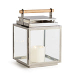 The Adwin Outdoor Lantern features a metal and glass design with a teak handle and includes a glowing white pillar candle inside.