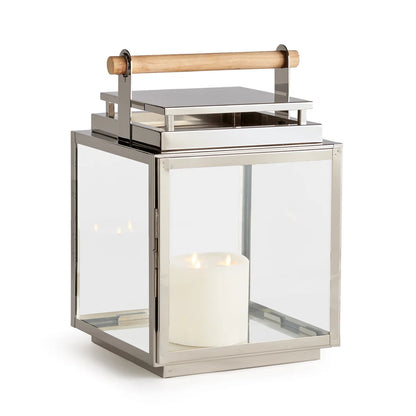 The Adwin Outdoor Lantern features a metal and glass design with a teak handle and includes a glowing white pillar candle inside.