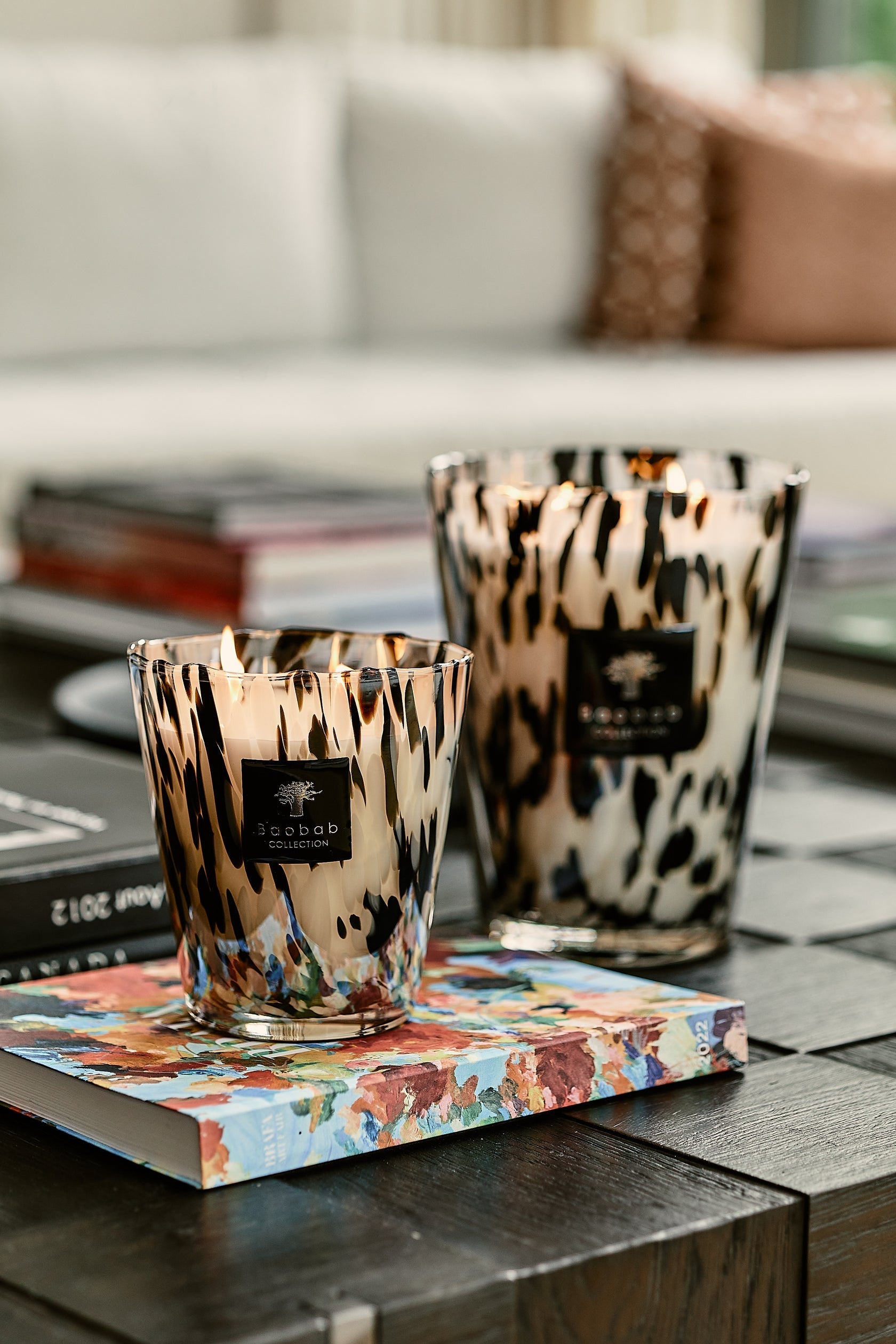 Two Baobab Collection Pearls Black Candles are placed on a book on a wooden table, with a sofa in the background.