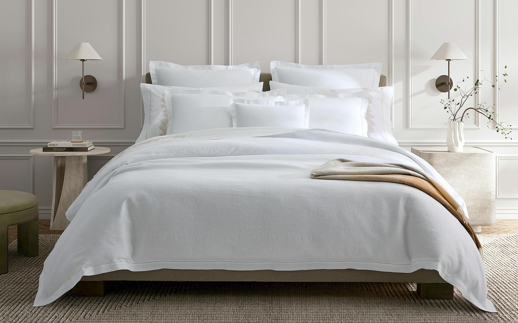 A neatly made bed with the Matouk Roman Linen Hemstitch Bedding Collection, White, pillows, and a beige throw blanket features beautiful hemstitch detailing. The room includes a white paneled wall, wall-mounted sconces, and a nightstand with a glass of water and a small plant.