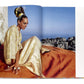 A woman in Caftans: From Classical to Camp sits outdoors on an orange fabric, with rocky terrain and a clear blue sky in the background.