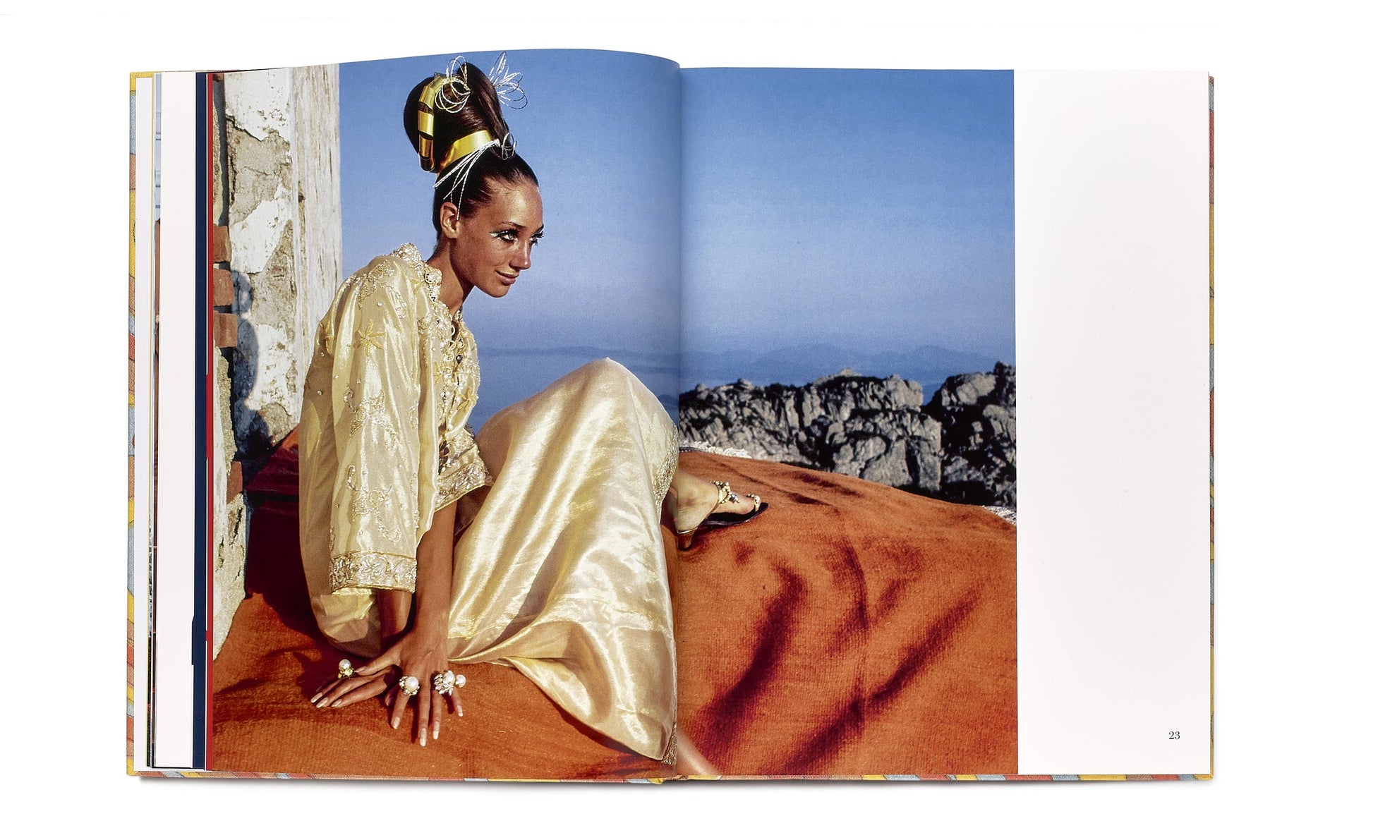 A woman in Caftans: From Classical to Camp sits outdoors on an orange fabric, with rocky terrain and a clear blue sky in the background.