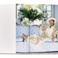 A woman in a light-colored caftan dress and hat reclines on a blue-patterned sofa, with her arm resting on the back. She is barefoot, and there's a leafy plant in the background. The scene evokes the elegance of Caftans: From Classical to Camp and is photographed in a book.