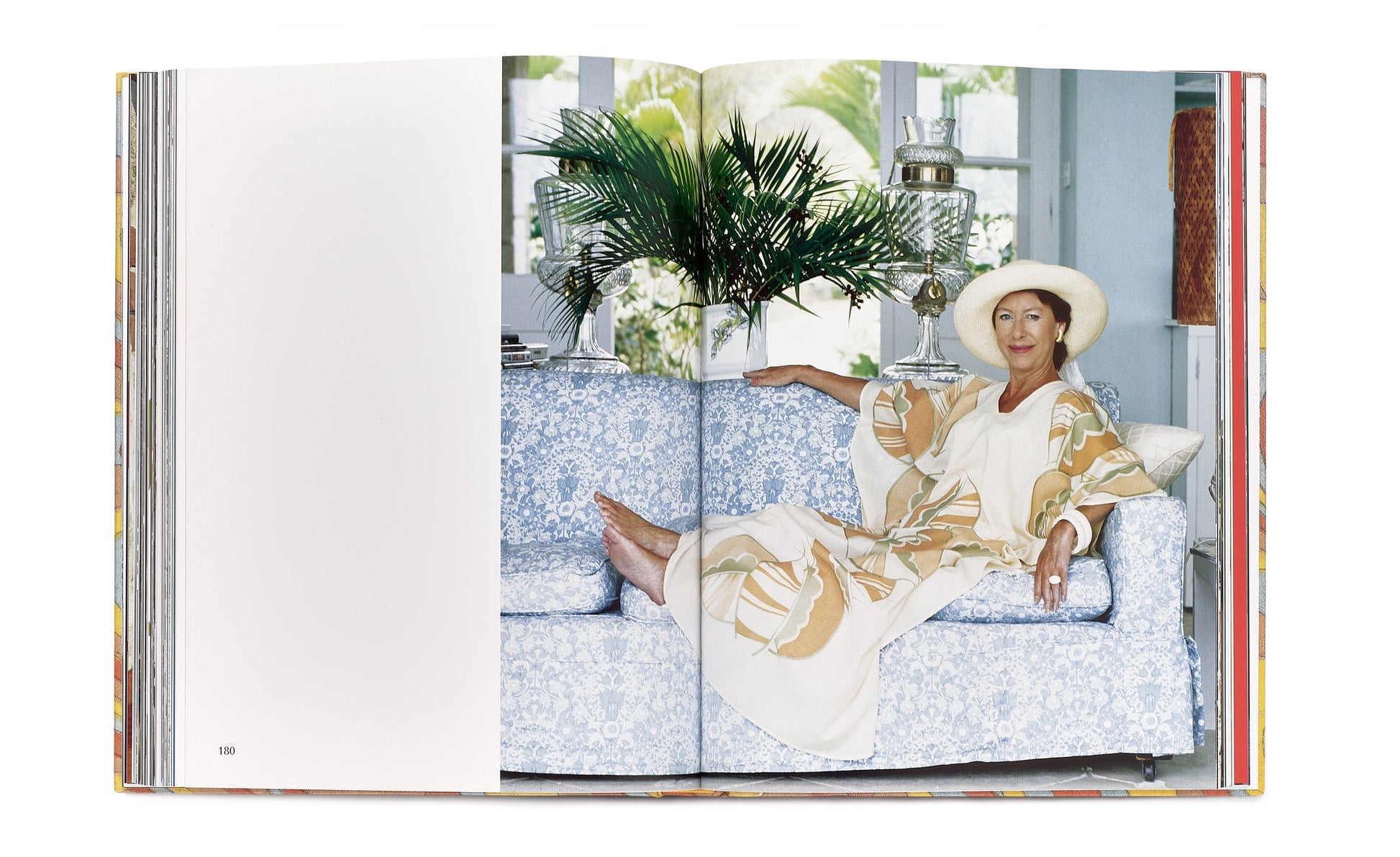 A woman in a light-colored caftan dress and hat reclines on a blue-patterned sofa, with her arm resting on the back. She is barefoot, and there's a leafy plant in the background. The scene evokes the elegance of Caftans: From Classical to Camp and is photographed in a book.