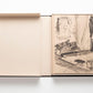 An open book box revealing an illustrated page inside. The left side is empty, with a beige cover reminiscent of Chanel's iconic looks, and the right side shows a black and white drawing with handwritten text from *The Impossible Collection: Chanel*.