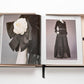 Open book displaying images of elegant black clothing with a white flower accessory on the left page and a long, lace-trimmed black dress on the right page, placed in a light-colored box with a black ribbon. This showcase is the epitome of luxury fashion, reminiscent of The Impossible Collection: Chanel's iconic looks.