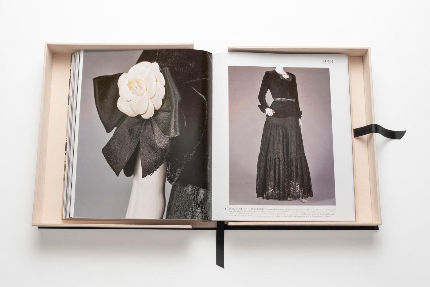 Open book displaying images of elegant black clothing with a white flower accessory on the left page and a long, lace-trimmed black dress on the right page, placed in a light-colored box with a black ribbon. This showcase is the epitome of luxury fashion, reminiscent of The Impossible Collection: Chanel's iconic looks.