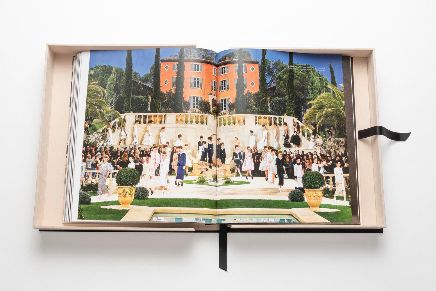 An open copy of The Impossible Collection: Chanel featuring an elaborate outdoor fashion show with models showcasing iconic looks on a tiered stage in front of a large, orange building surrounded by greenery.