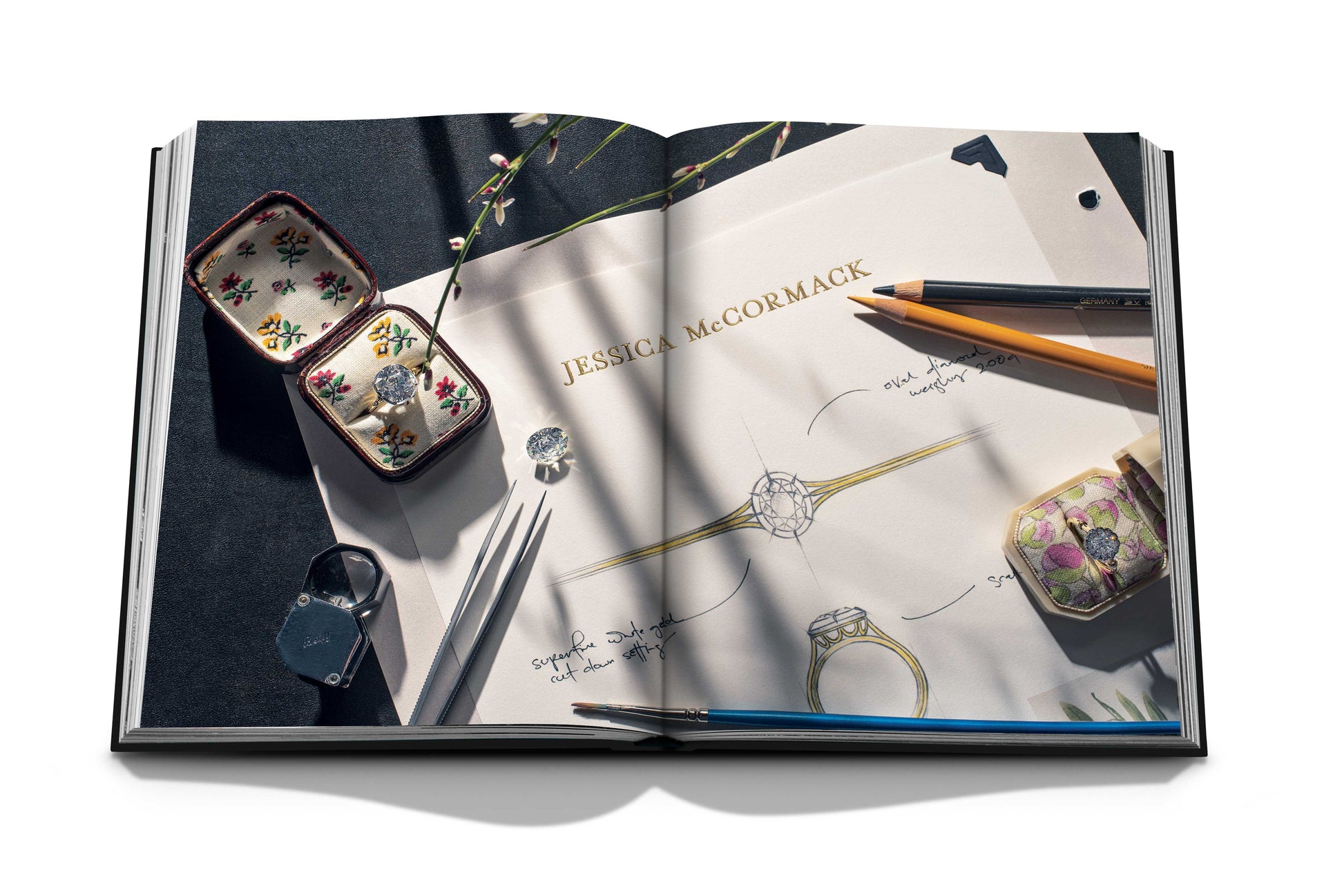 An open book displays a jewelry design sketch titled "Diamonds: Diamond Stories," surrounded by rings crafted from captivating stones, pencils, and a ring box on a dark surface.